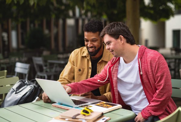 Two men sit at an outdoor table, smiling and looking at a laptop screen. One is dressed in a yellow jacket, and the other in a red hoodie. They are discussing the Support Coordination Pricing Freeze.