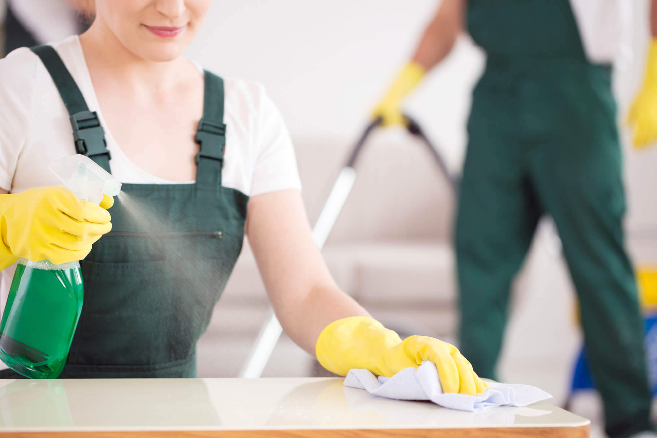 Two people in green overalls and yellow gloves are cleaning. One person in the foreground is wiping a surface with a cloth and a spray bottle, while the other vacuums in the background.