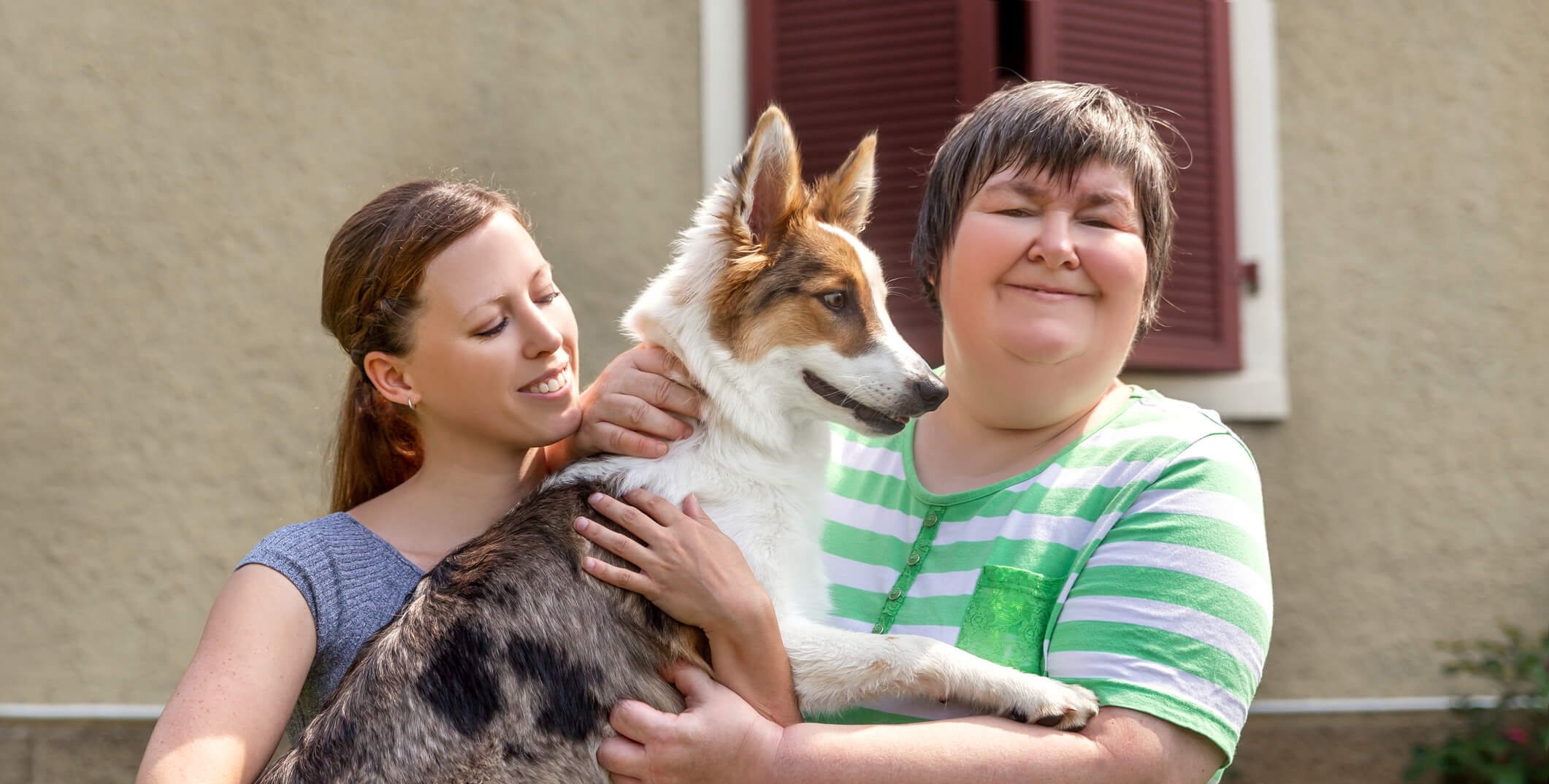 Two women smiling and holding a dog together outdoors, representing companionship and supportive psychosocial coaching relationships.